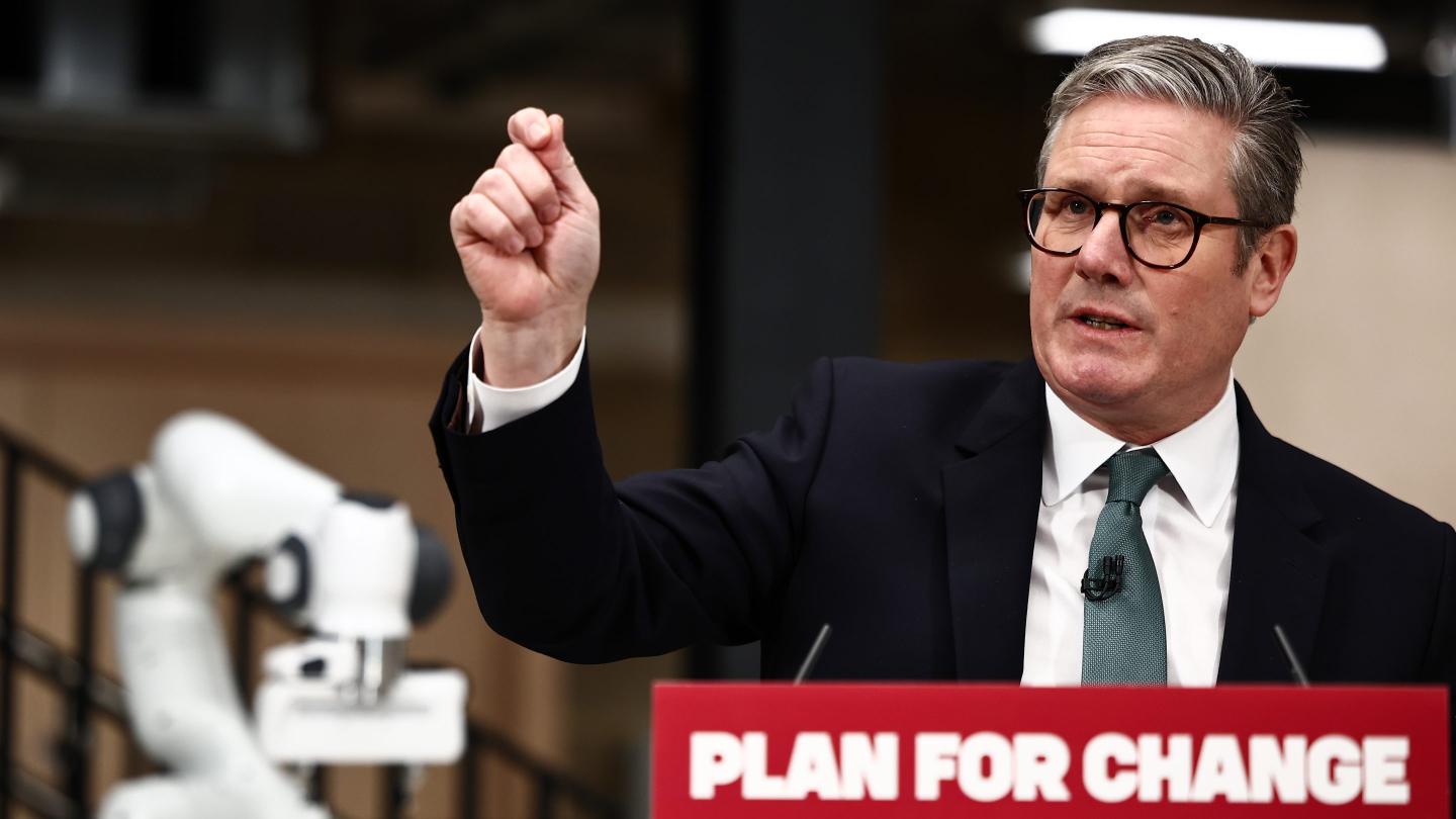 U.K. Prime Minister Keir Starmer speaking before a podium with a sign saying "Plan for Change" on it and holding his right arm up in a gesture, while a robot arm is visible in the background.