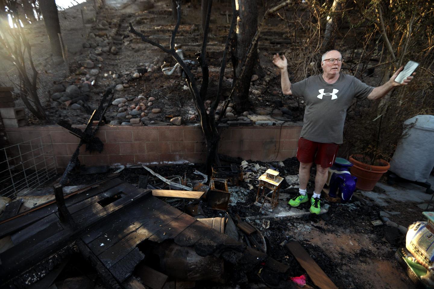 A bald overweight white man wearing an Under Armour shirt and shorts holds his hands up in disbelief as he stands in the wreckage of his former home, a result of the devastating Los Angeles wildfires