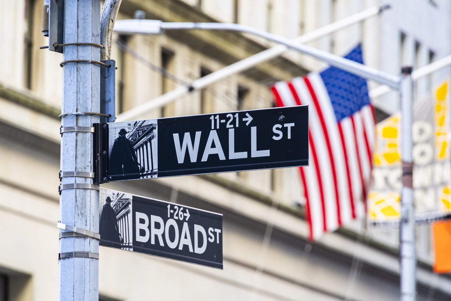 Wall Street sign inscription as seen with the American flag in the background on the road and buildings of Wall St.
