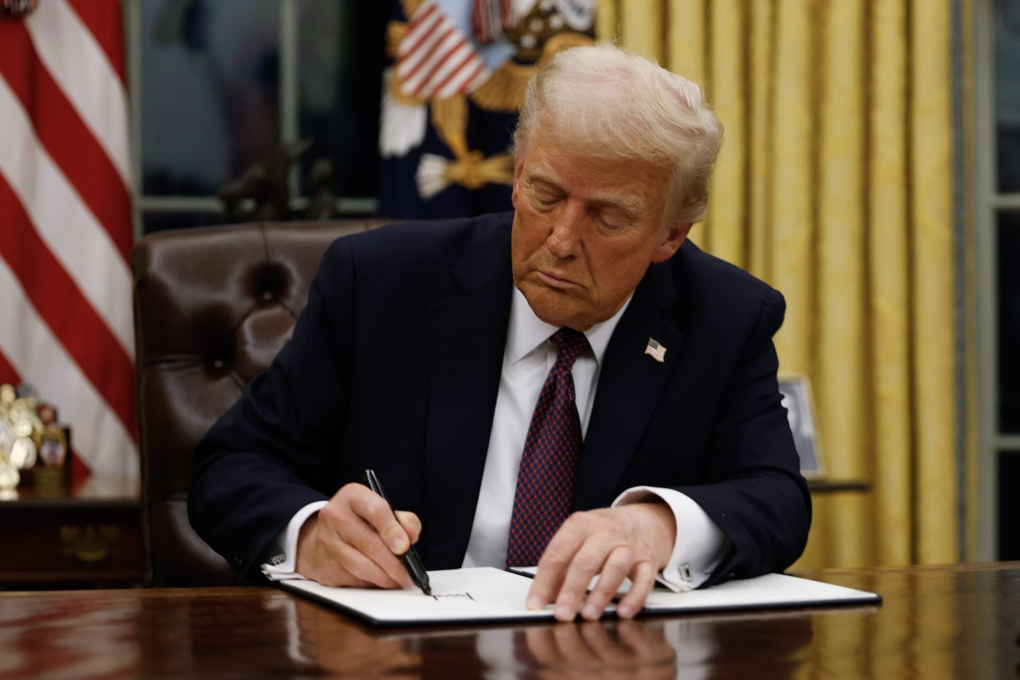 Donald Trump sits at his desk and signs a document.