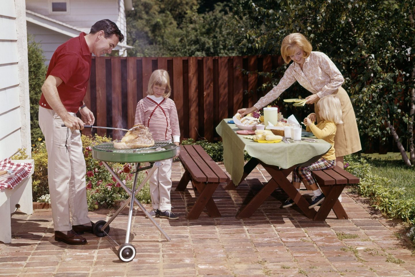 A man grills meat in his backyard while a young girl looks on. Next to them, a woman sets a picnic table.