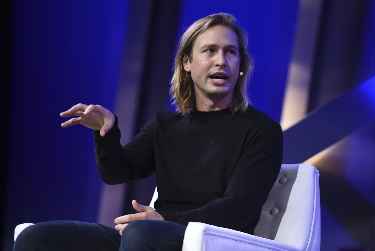 man with shoulder length blonde hair wearing a black sweater, speaking onstage