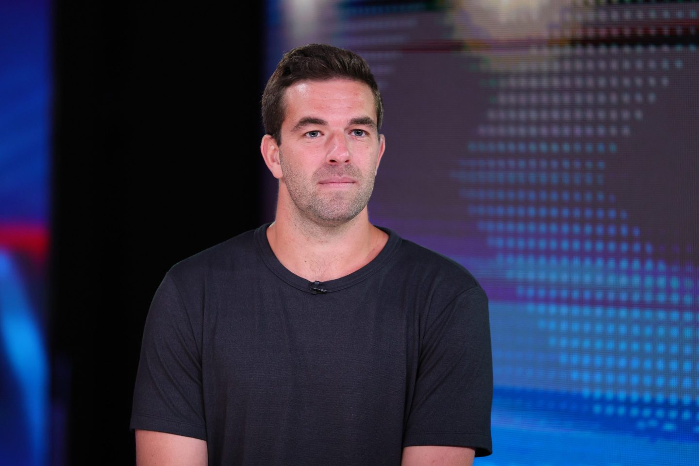 Billy McFarland, in a black T-shirts, sits in front of a patterned blue background.