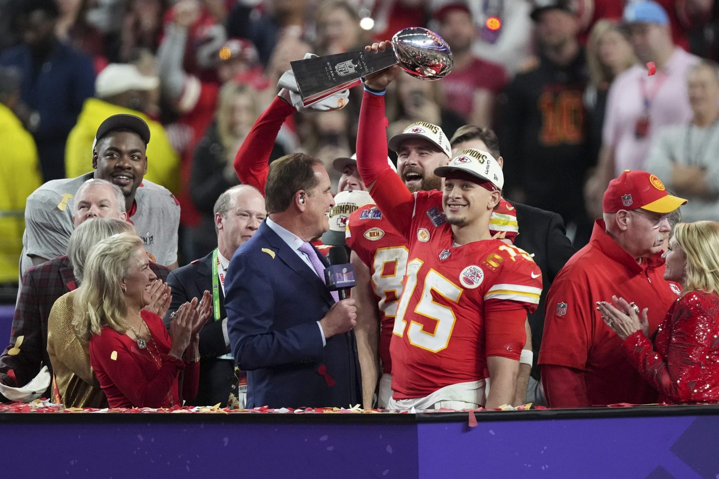 Kansas City Chiefs QB Patrick Mahomes raises the Vince Lombardi Trophy toward the crowd as he and teammates are interviewed on a platform.