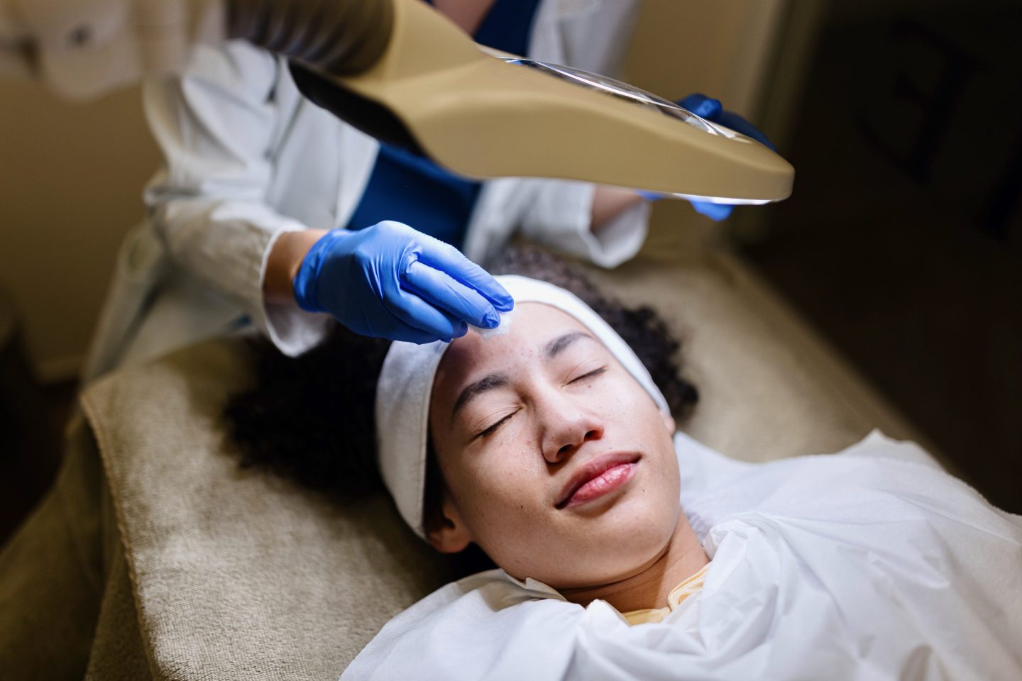 young girl's face being examined under a light with dermatologist's gloved hand in the image