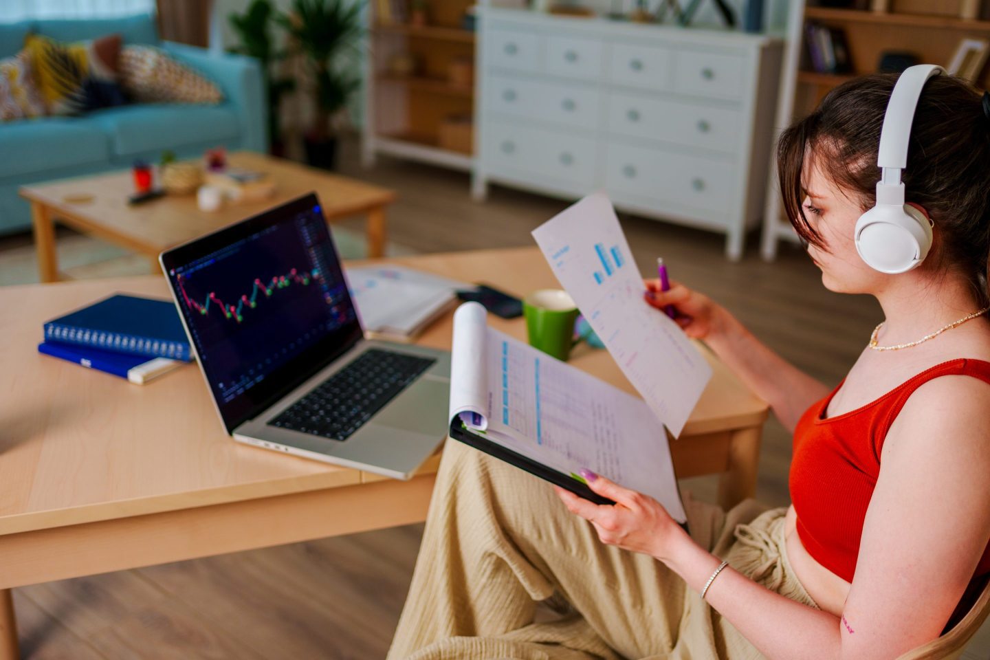 Young woman with headphones looks at a stock exchange price chart on a laptop, while holding documents in her hand.