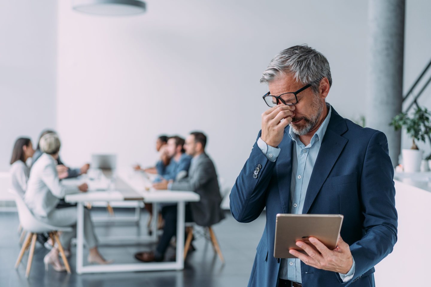 A businessman with a headache rubs his eyes as his colleagues work in the background.