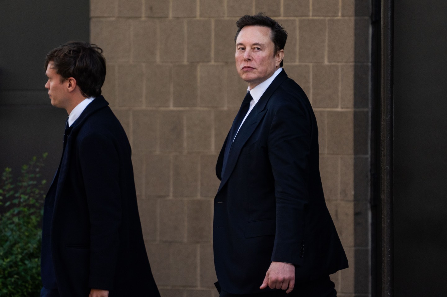 Elon Musk walking and wearing a black suit.