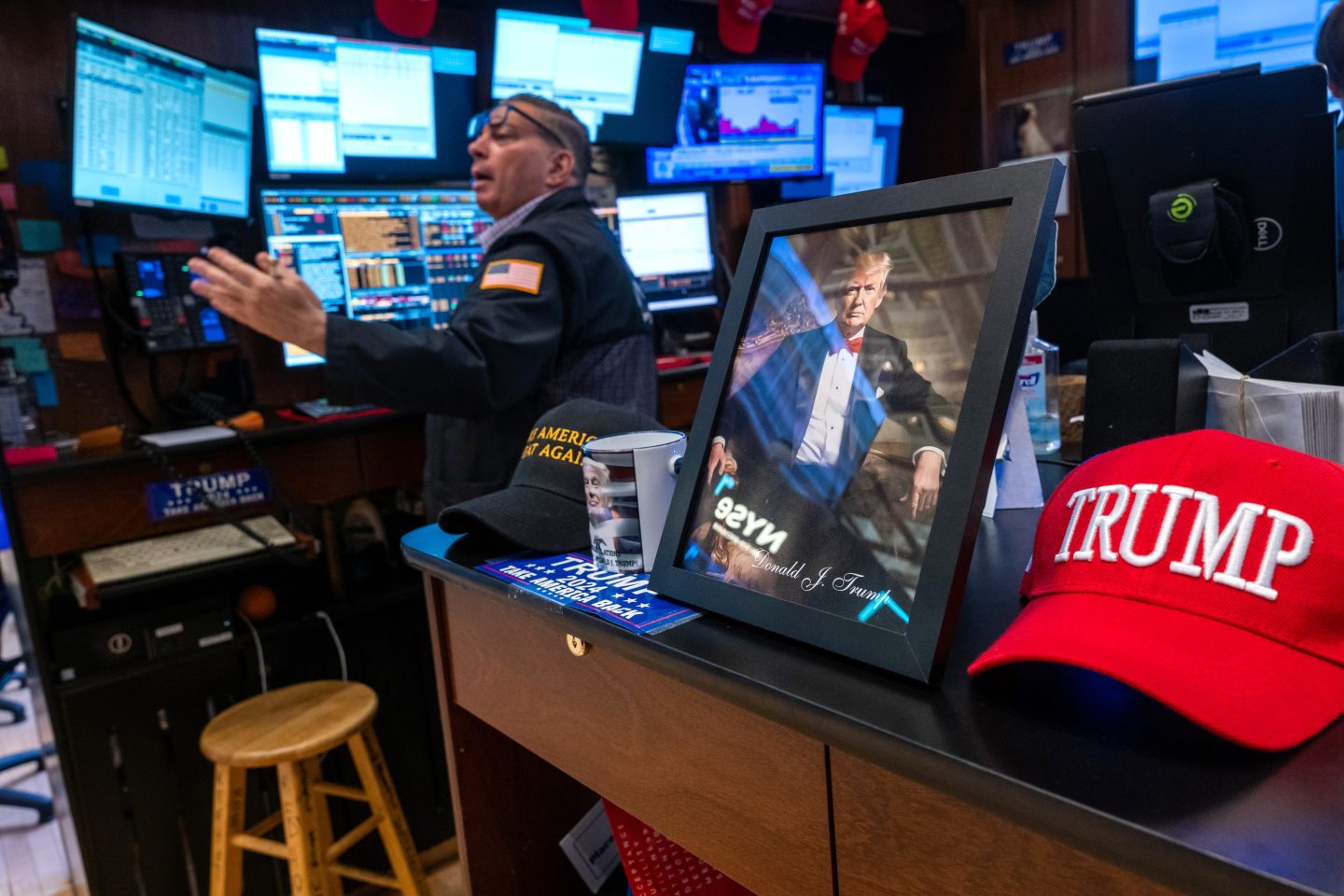 A trader looks to his left with a hand out and glasses perched above his eyes as he stands in front of his various screens. A picture of Donald Trump and other related memorabilia sits in the foreground.