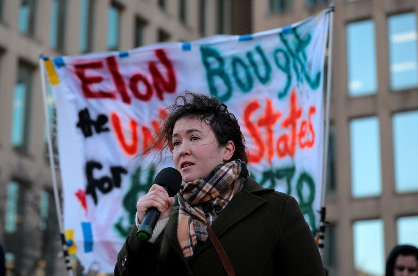 A person with a microphone stands in front of a protest sign.