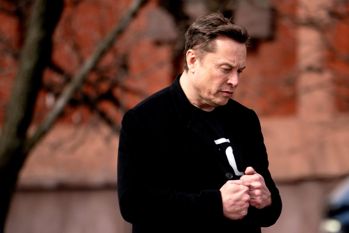 Elon Musk, wearing a black jacket and standing outside, looks down with his hands clasped.