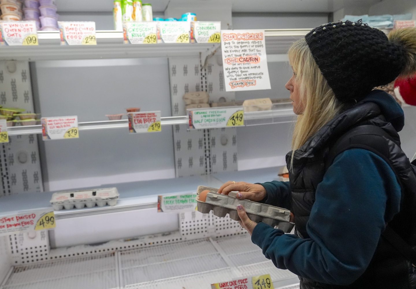 A woman holds a carton of eggs and looks at an empty refrigerated shelf at a grocery store.