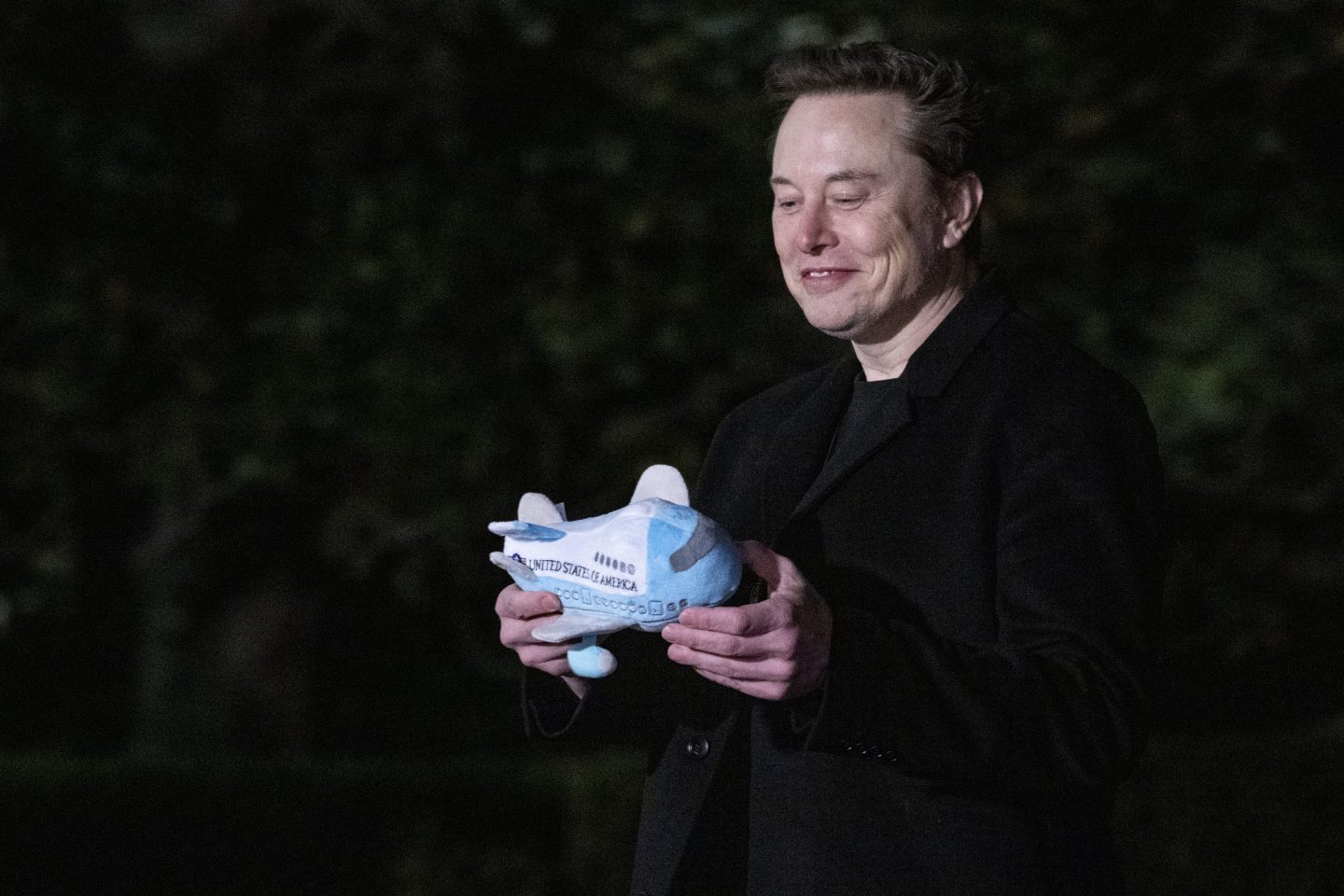 Elon Musk looks down and smiles at a small stuffed felt airplane.