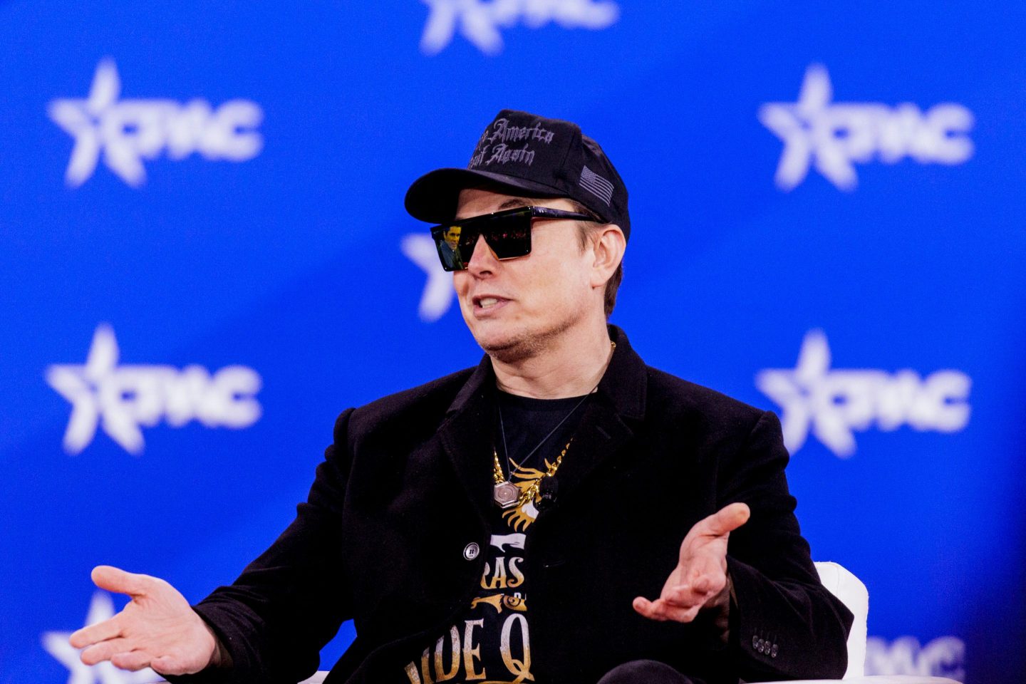 Elon Muks, in sunglasses and a black MAGA hat, shrugs as he sits in front of a blue CPAC background.