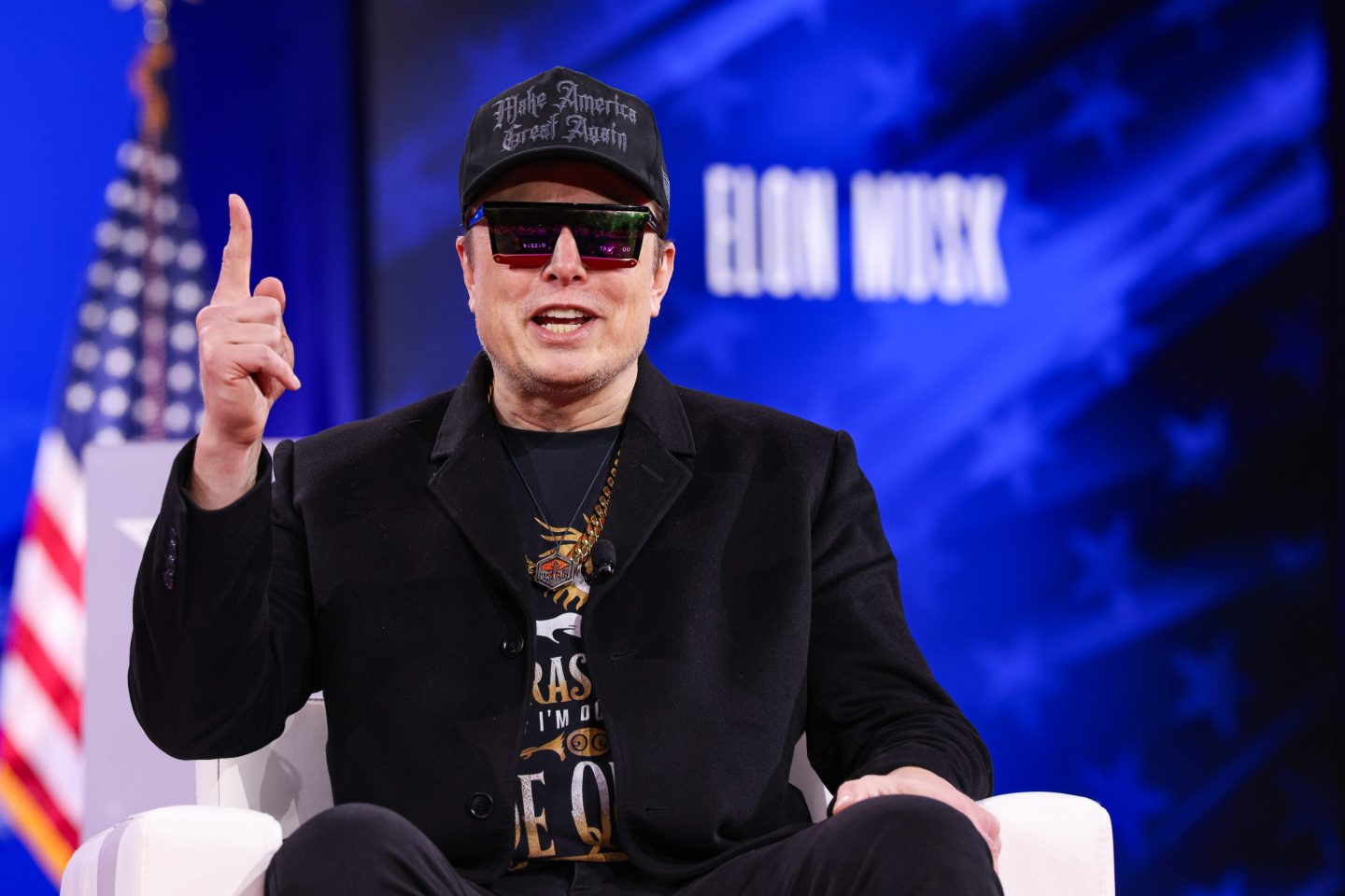 Elon Musk, sitting in a chair wearing all black and black sunglasses, lifts his finger while speaking