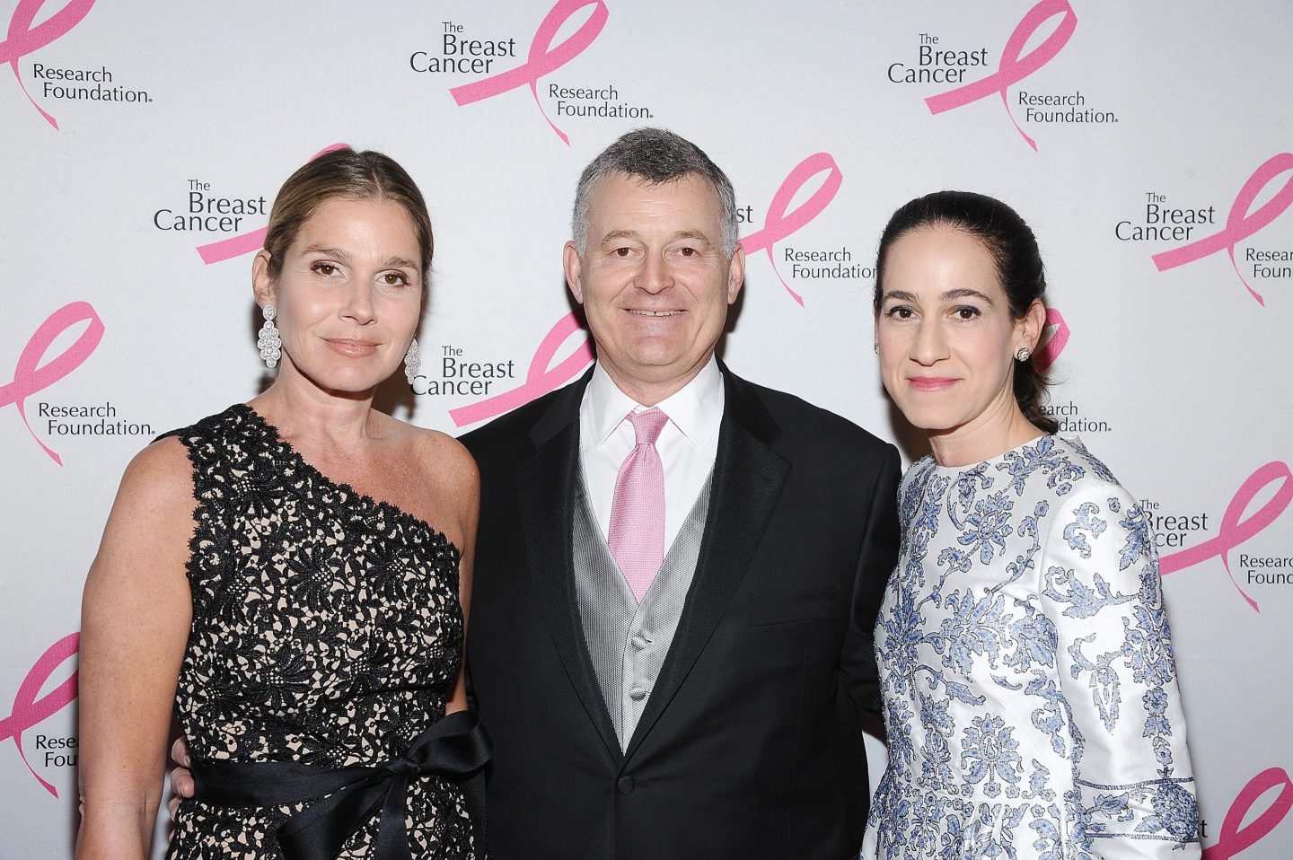 Two women stand at the side of one man at a red carpet event.