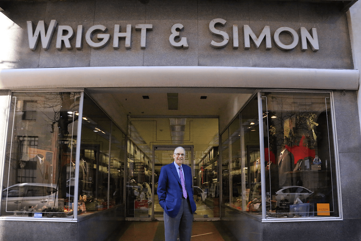 Wright & Simon owner Leonard Simon, dressed in dark pants, a blue shirt, dark blue blazer, and purple tie, stands in front of his store.
