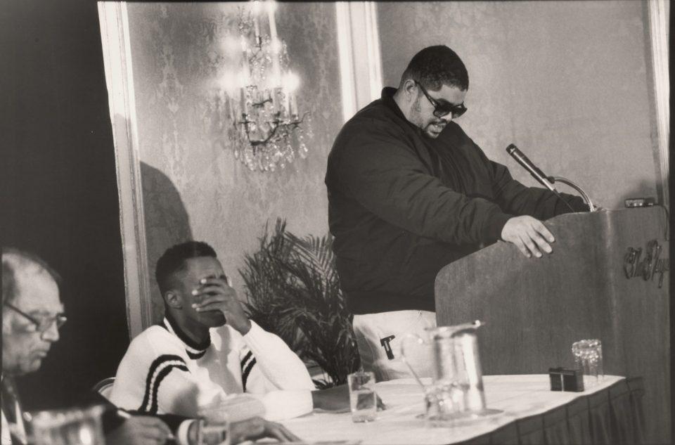 Sean Combs sits with his head in his hand as Heavy D stands at a podium with his head down.