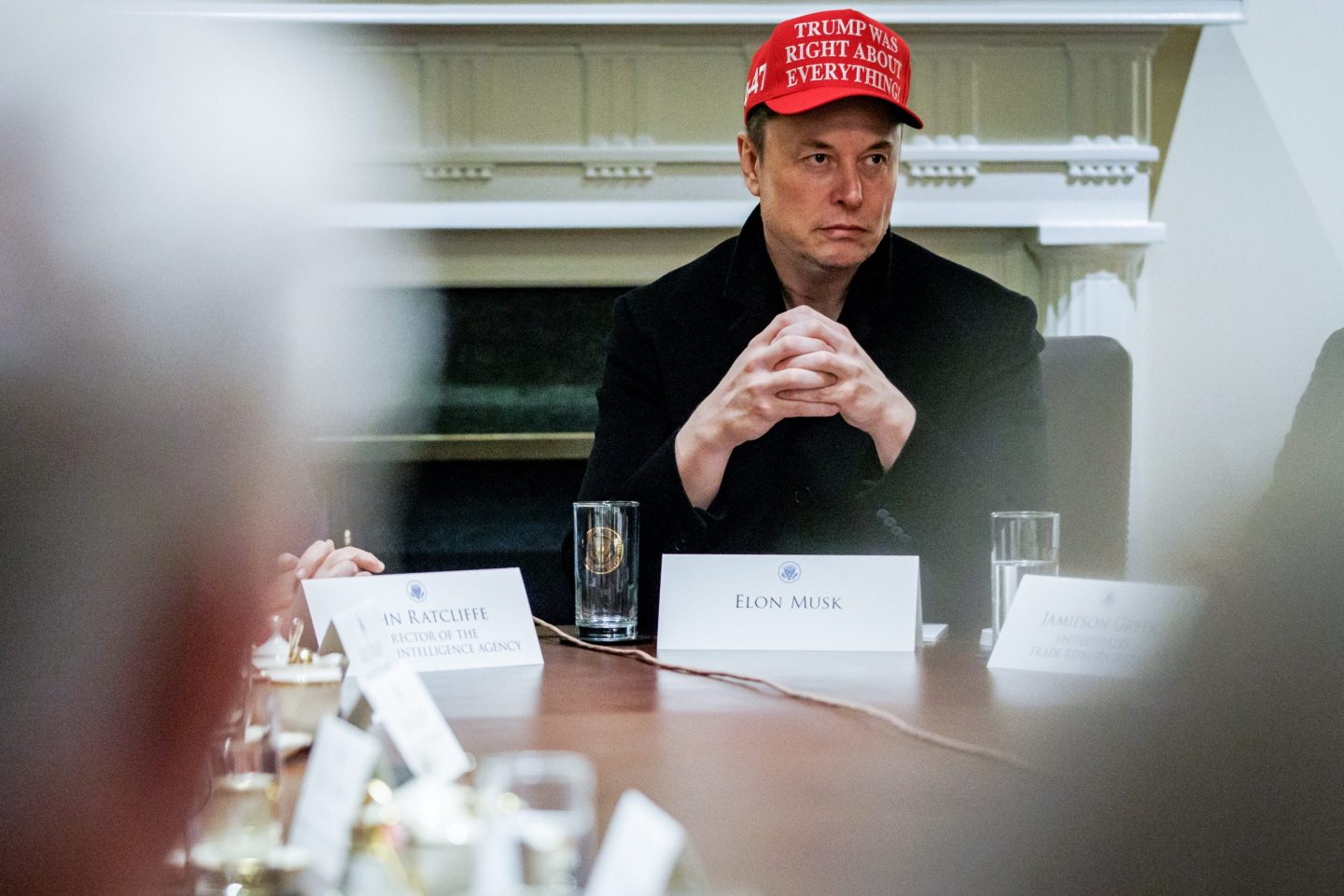 Elon Musk sits in a White House meeting wearing a red hat