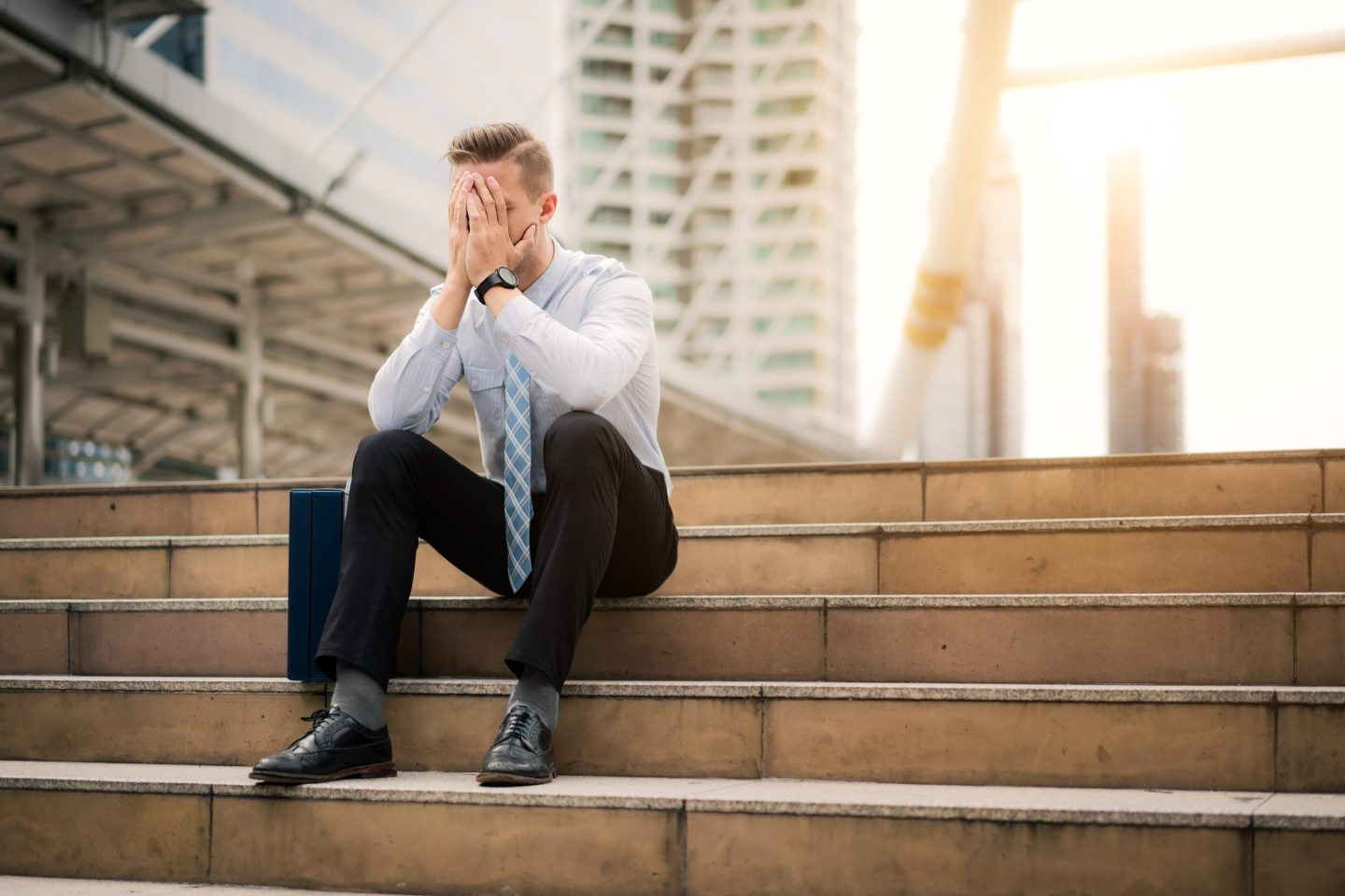 A young business man sits on steps and holds his head in his hands.