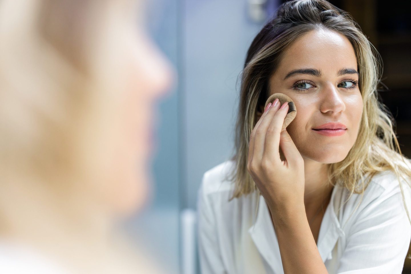 Reflection in a mirror of smiling woman applying face powder in a bathroom.