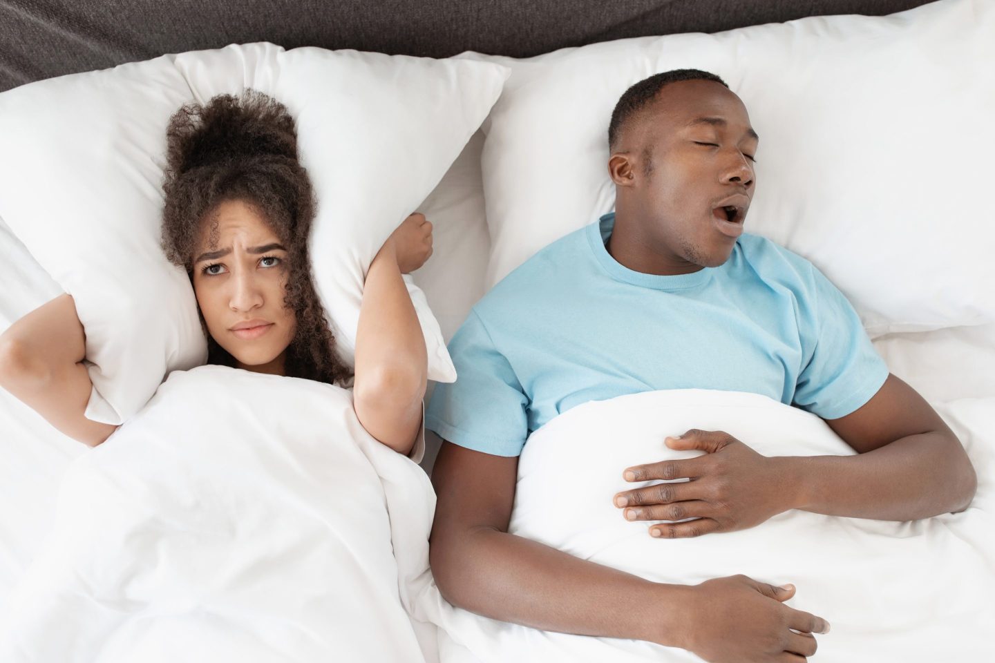 Young couple in bed, the woman awake and blocking her ears with a pillow, the man seemingly snoring.