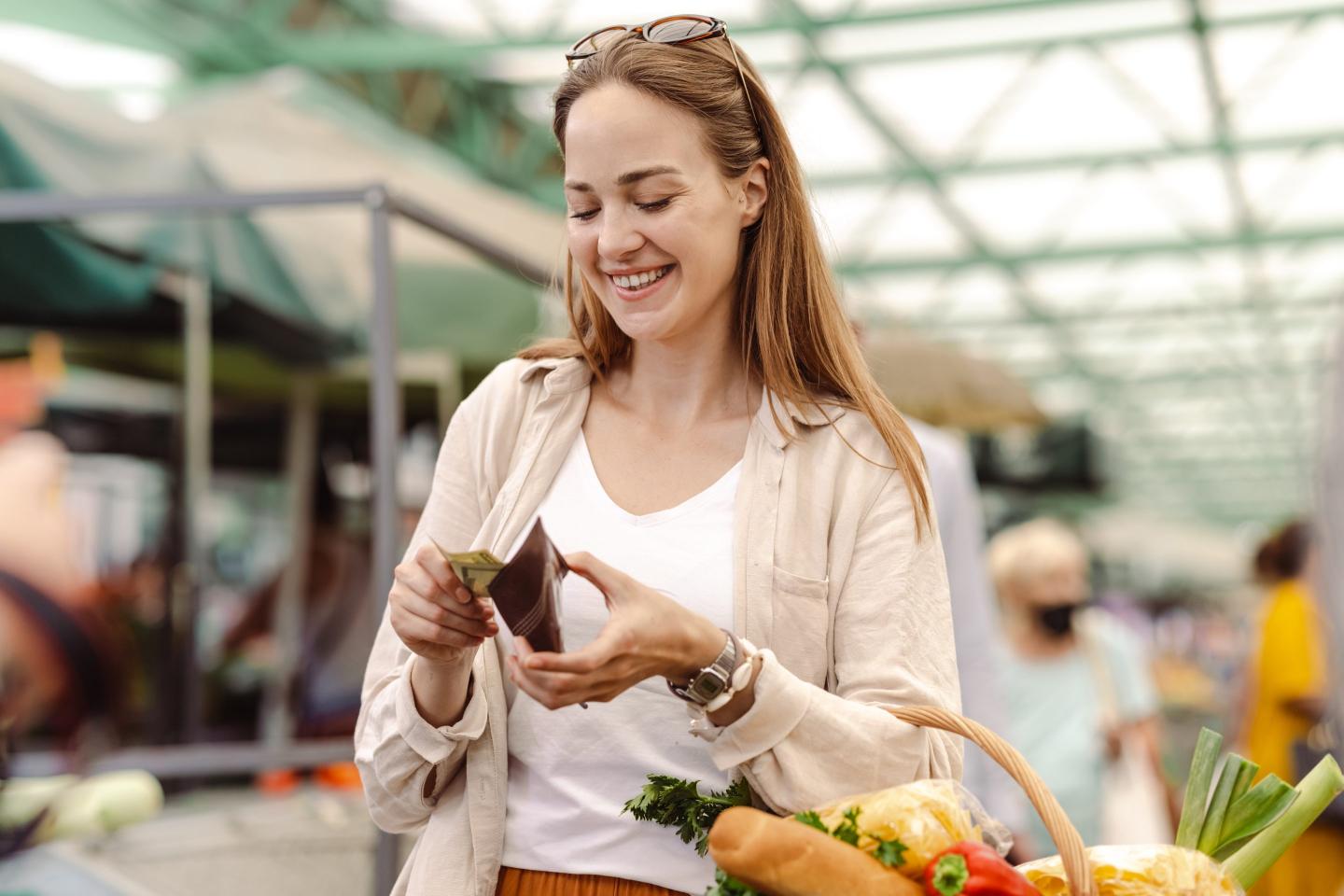 A smiling young woman is holding a basket full of groceries and counting money