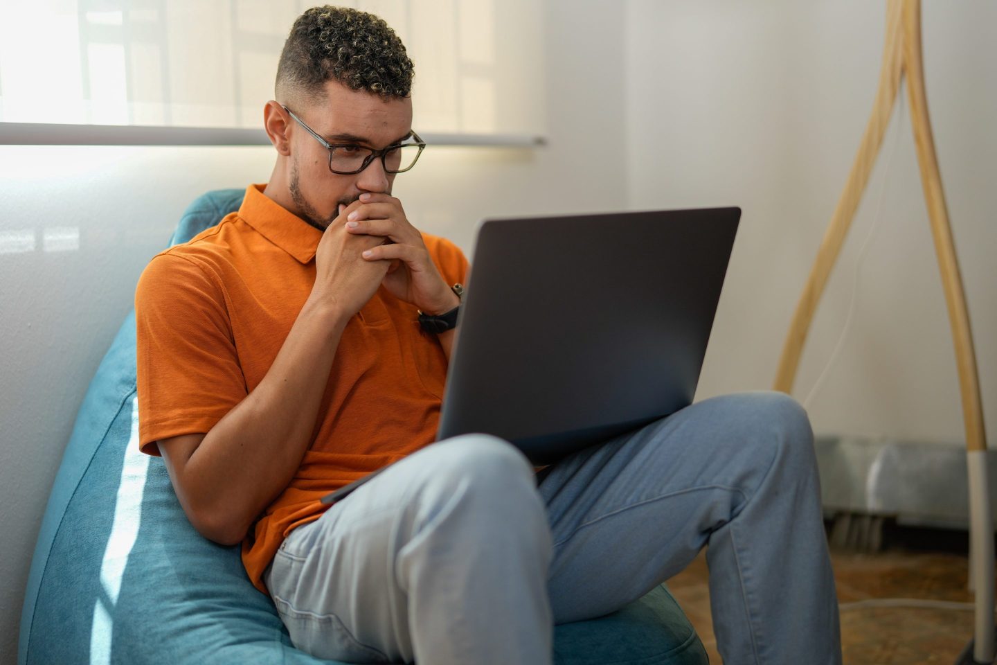 A man in a beanbag chair sits with his hands clasped over his mouth, with his laptop on his lap.