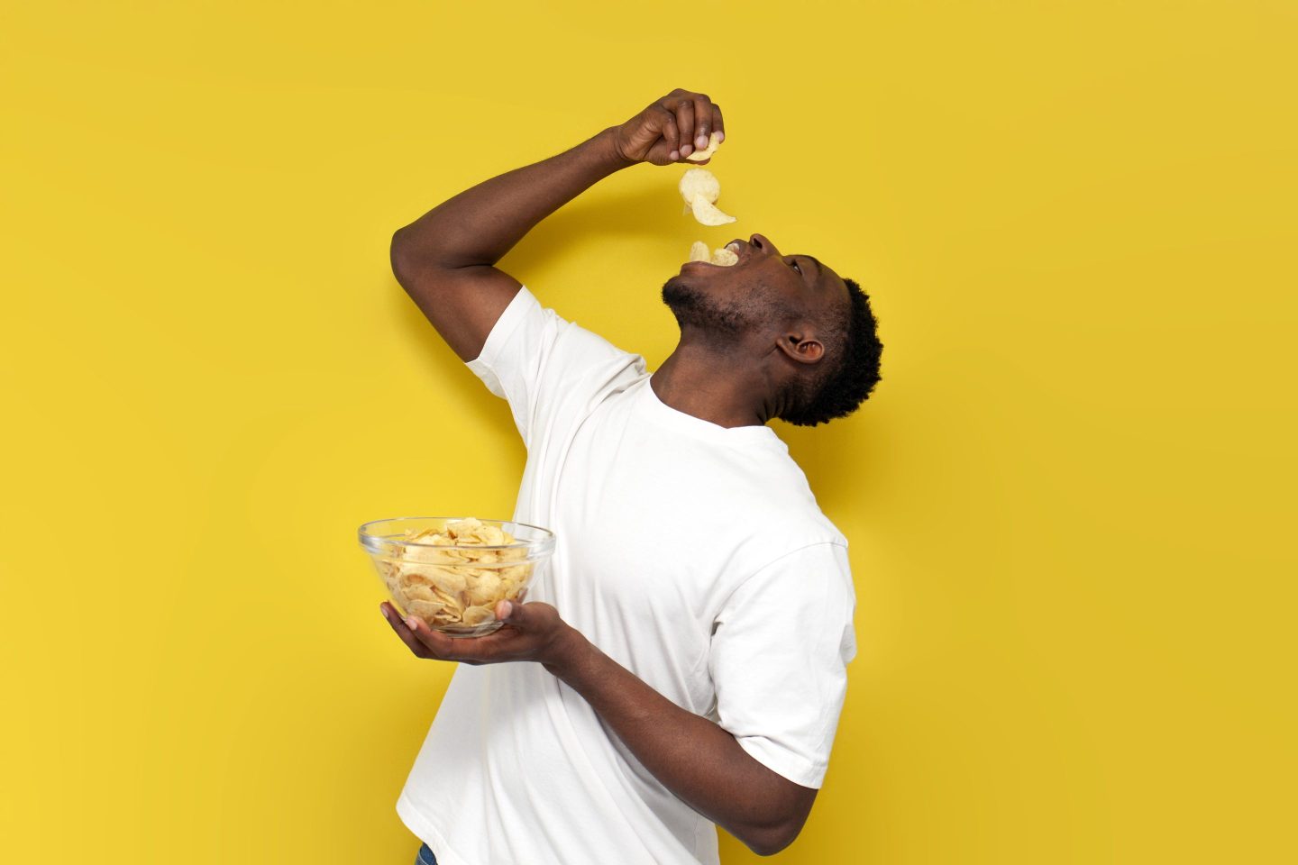 happy african american man holding plate of chips and eating snacks from potatoes on yellow isolated background, guy in white t-shirt eats fast food