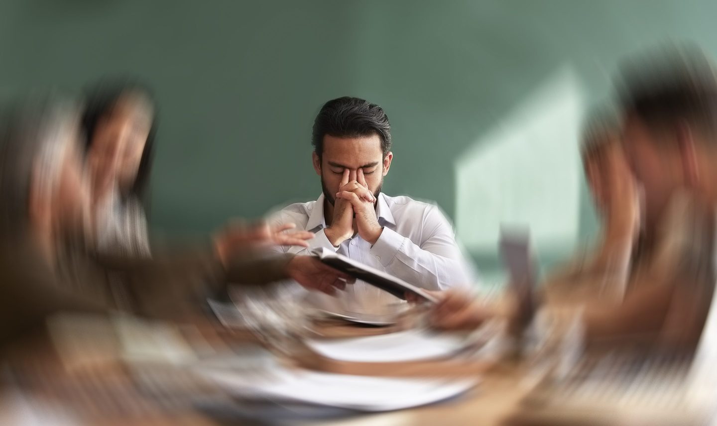 Overwhelmed employee in a meeting with his eyes closed and hands up to his temples.