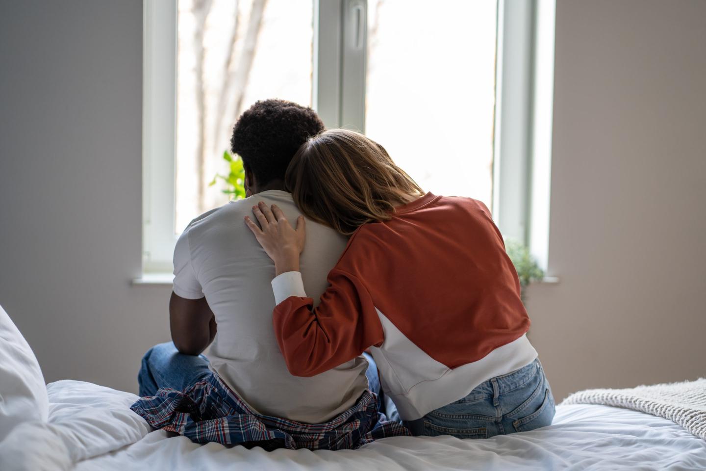 Two adults, seen from behind, sitting on a bed and leaning into each other in front of a window.