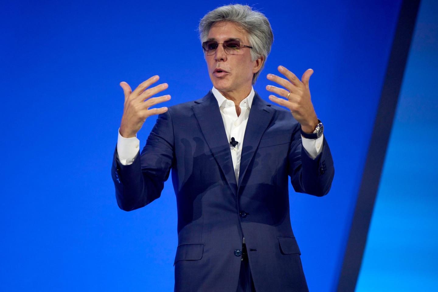 Bill McDermott, chief executive officer of ServiceNow Inc. on stage, in front of a blue background, gesturing with both hands.