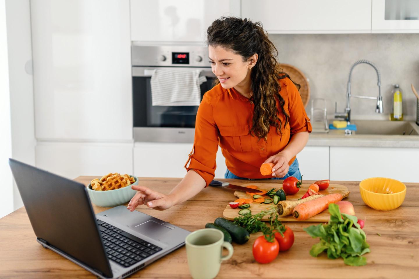 A woman prepares a meal in her kitchen while using a laptop