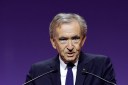 Bernard Arnault, wearing a dark suit, speaks into a microphone and is in front of a deep purple background.