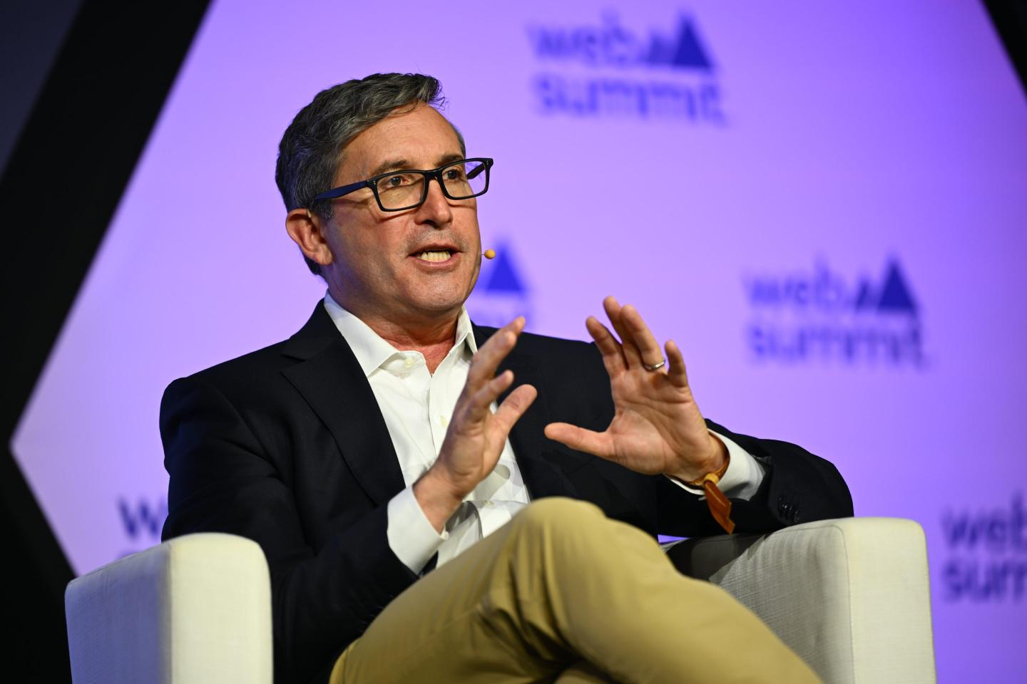 Man in white shirt, blazer and glasses sitting onstage at a conference and gesticulating with his hands