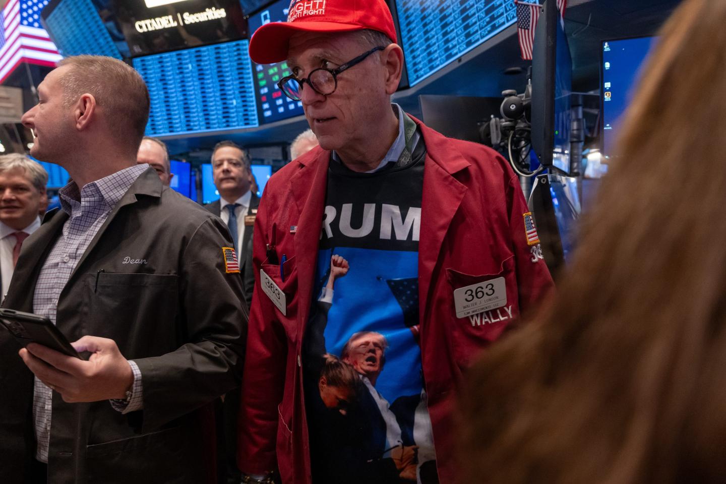A man wearing a Trump hat and shirt, along with his red trading uniform, looks to his right on the floor of the New York Stock Exchange.