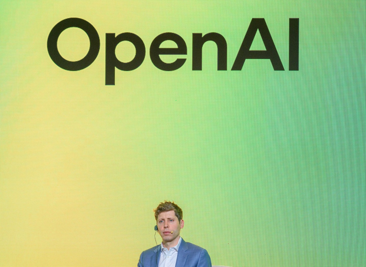 OpenAI CEO Sam Altman stands in front of a blue and yellow background on stage.