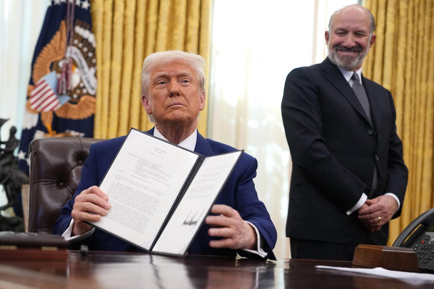 U.S. President holds up an executive order as Commerce Secretary Howard Lutnick smiles and looks on from the president's right.
