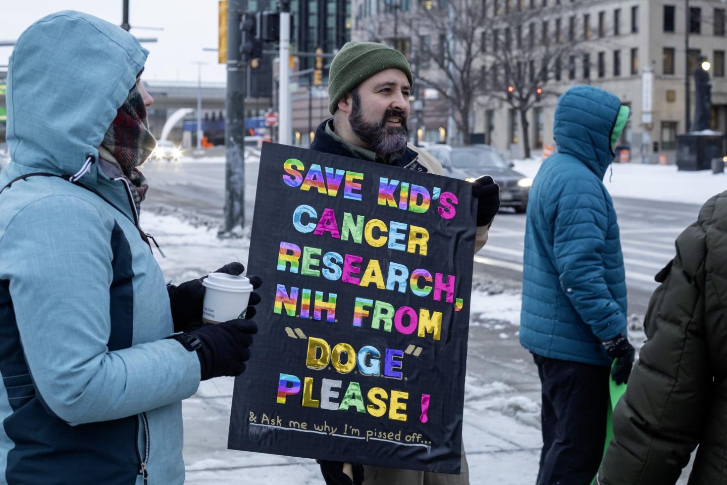 A man outside in the snow holds a sign with rainbow lettering that reads, "Save kid's cancer research from 'DOGE' please!"