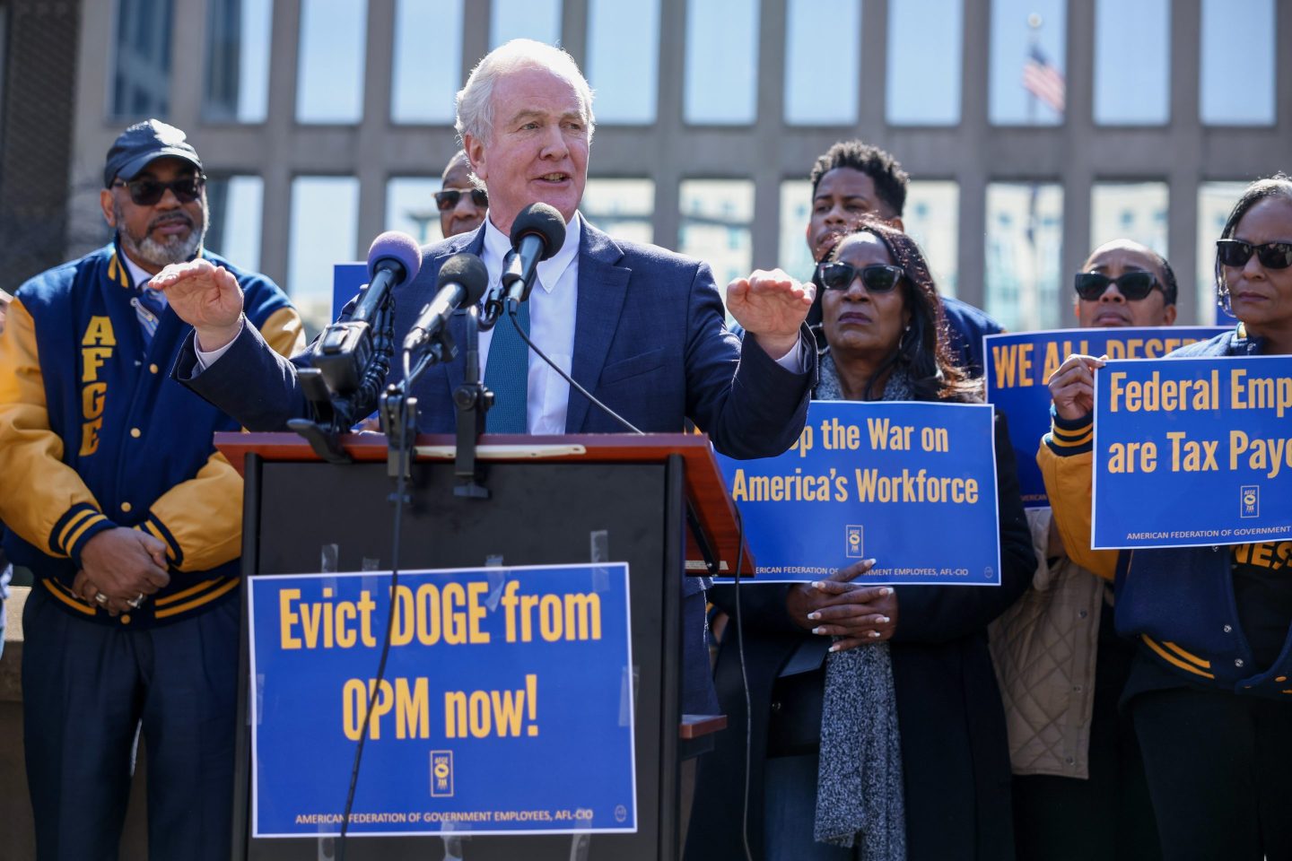 U.S. Sen. Chris Van Hollen speaks behind a podium with a blue and yellow sign that says, "Evict DOGE from OPM now!"