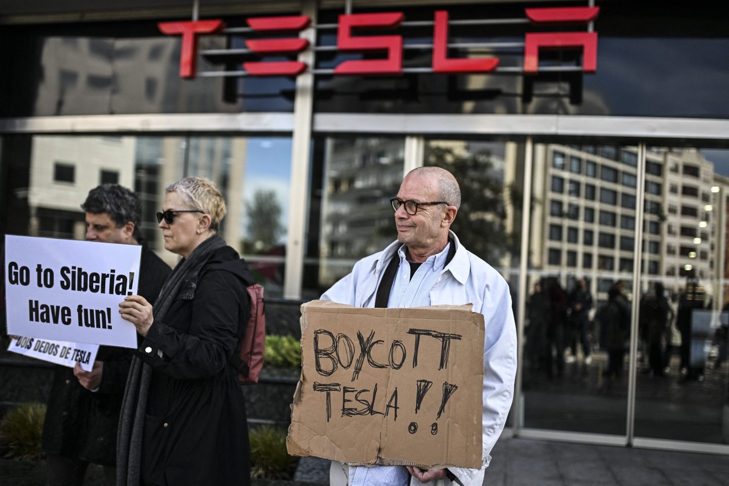A man holds a cardboard sign in front of a Tesla storefront that reads, "Boycott Tesla!!"