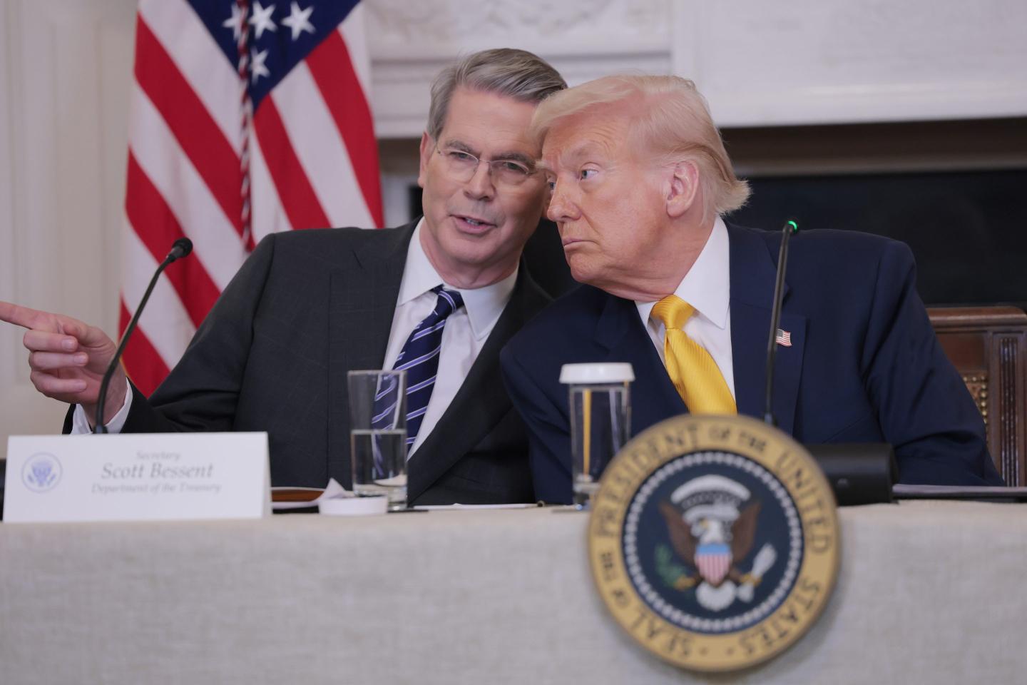 Donald Trump leans over to right, where Scott Bessent is sitting directly next to him and pointing with his right index finger in that direction.