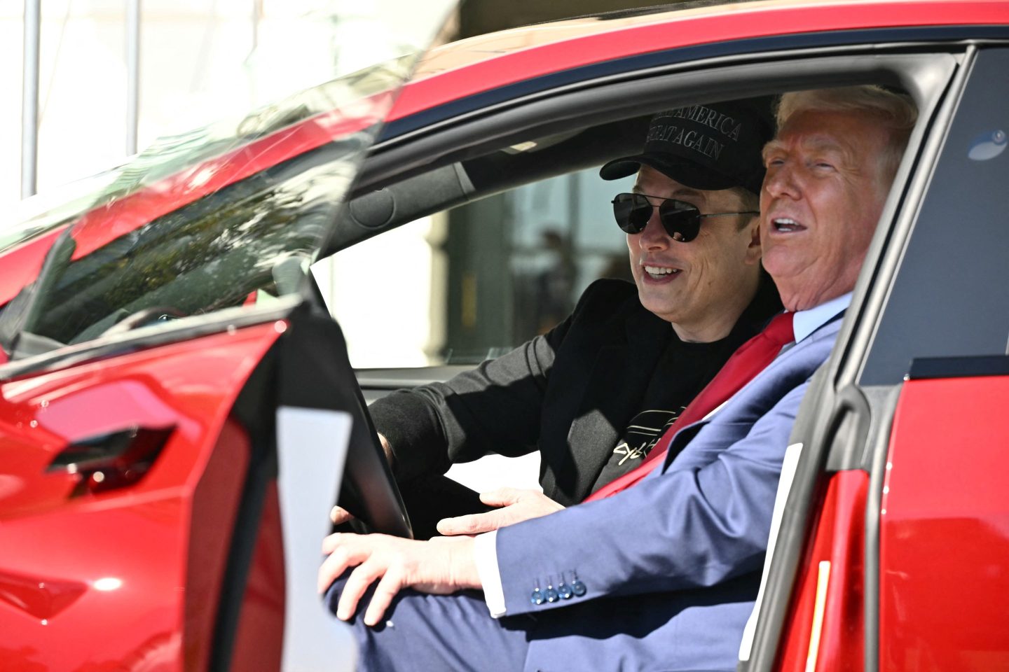 Elon Musk and Donald Trump sit in the front seats of a red Tesla, smiling.
