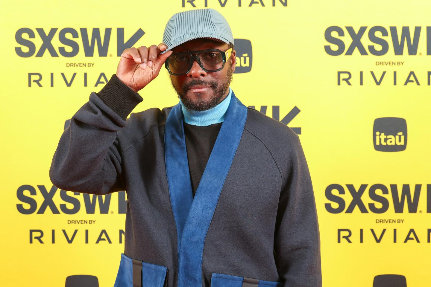 Will.i.am nods his hat standing behind a yellow SXSW background.