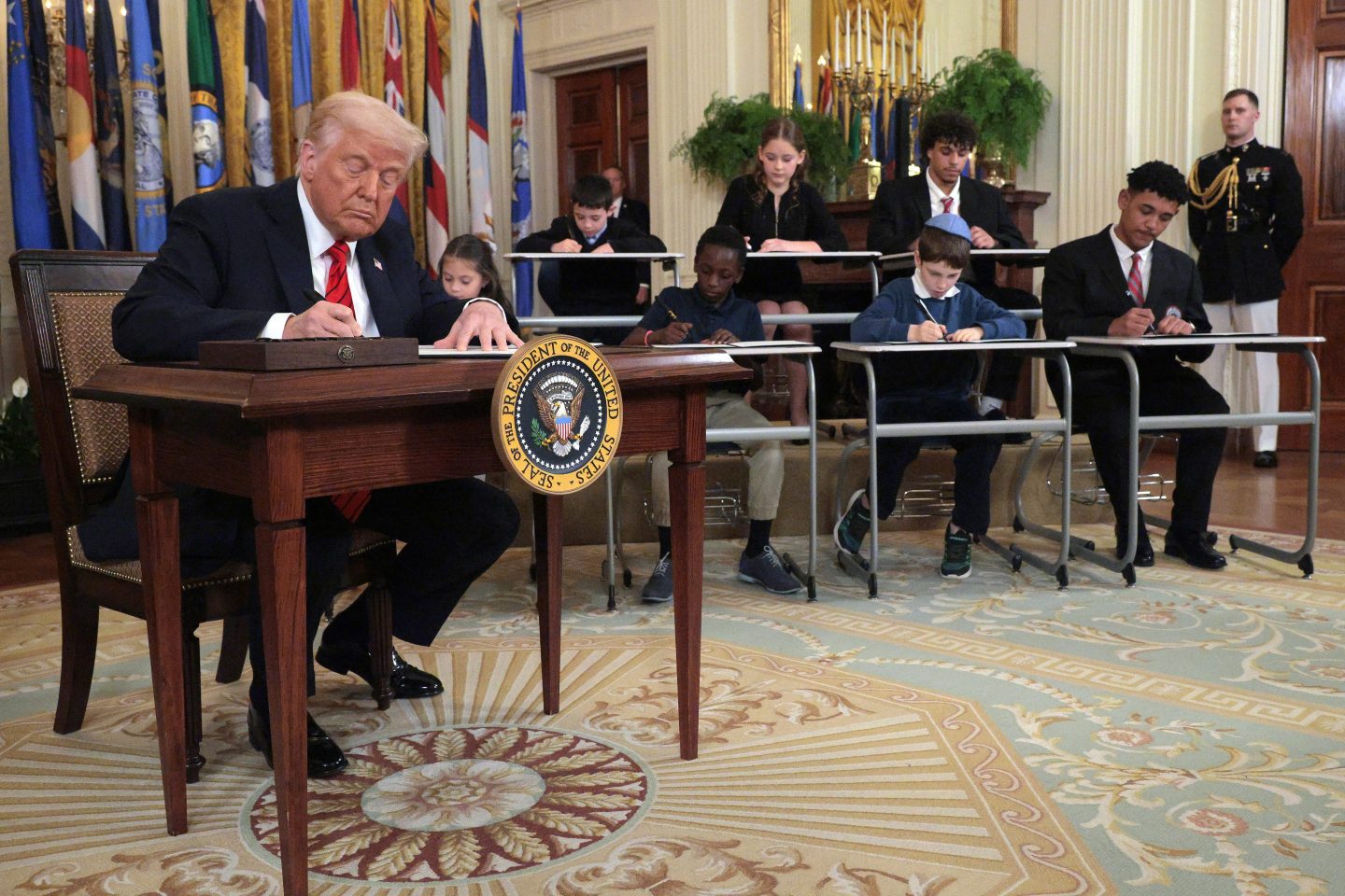 Trump sits at a desk flanked by schoolchildren