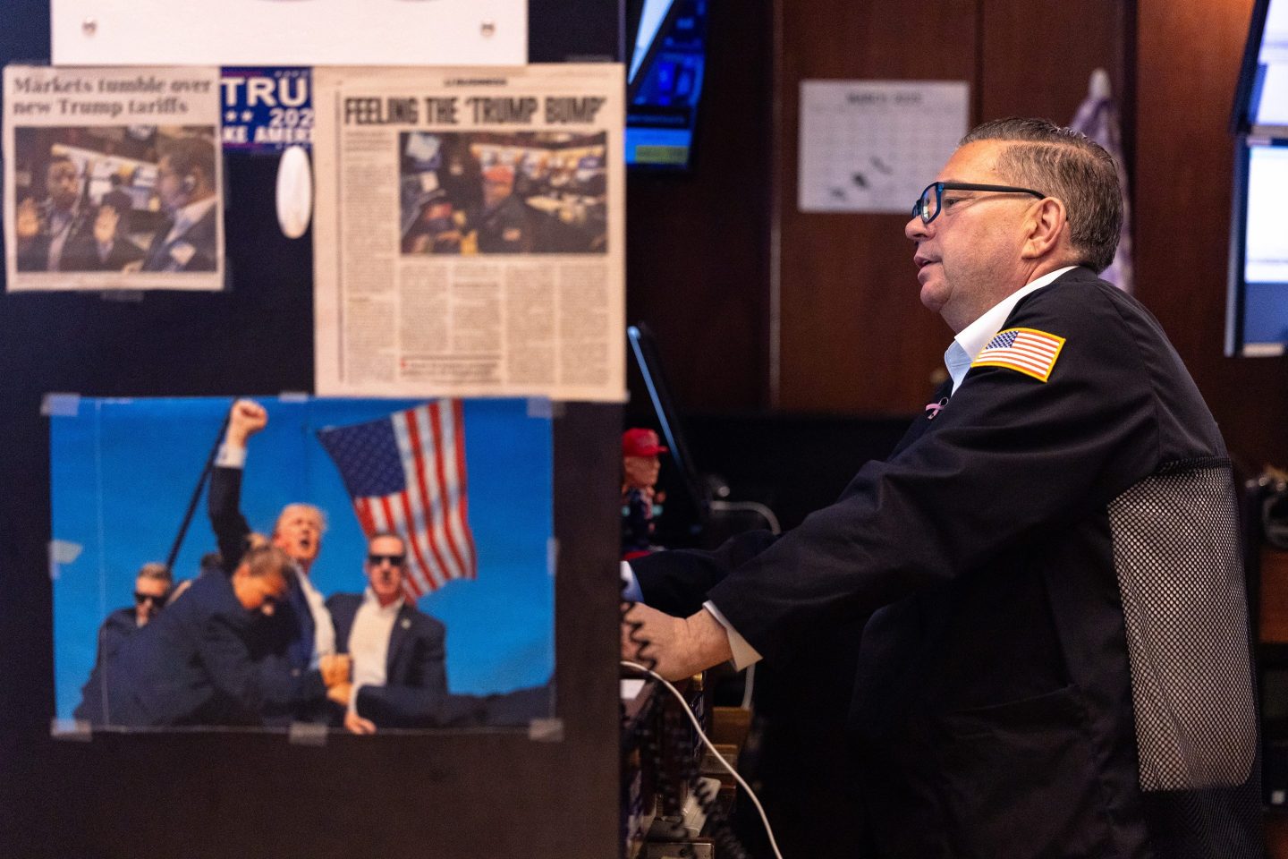 A trader on the floor of the New York Stock Exchange sits at his desk as pictures of President Donald Trump raising his fist after an assassination attempt and other news clippings are visible in the foreground.