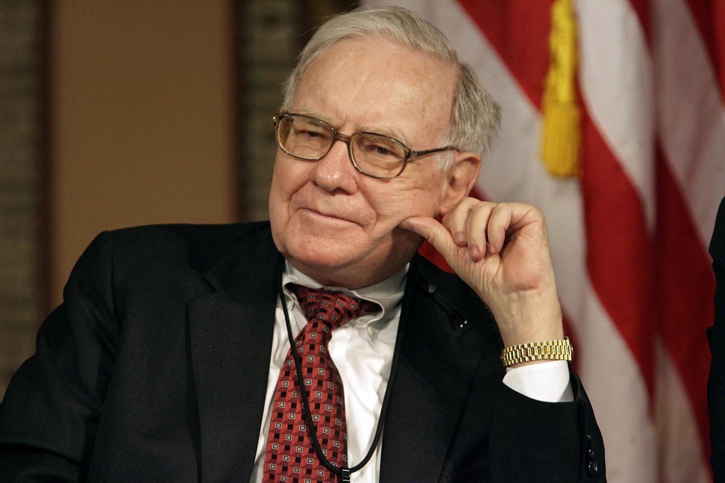 Warren Buffett sits and leans his head on his left hand as he listens with an American flag in the background.