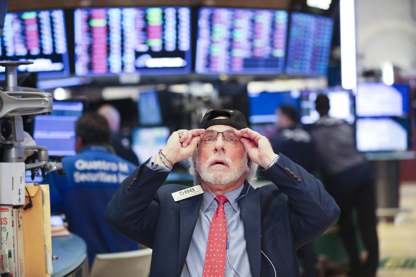 A man in a jacket and tie grabs his glasses and makes a very concerned face on the floor of the New York Stock Exchange.