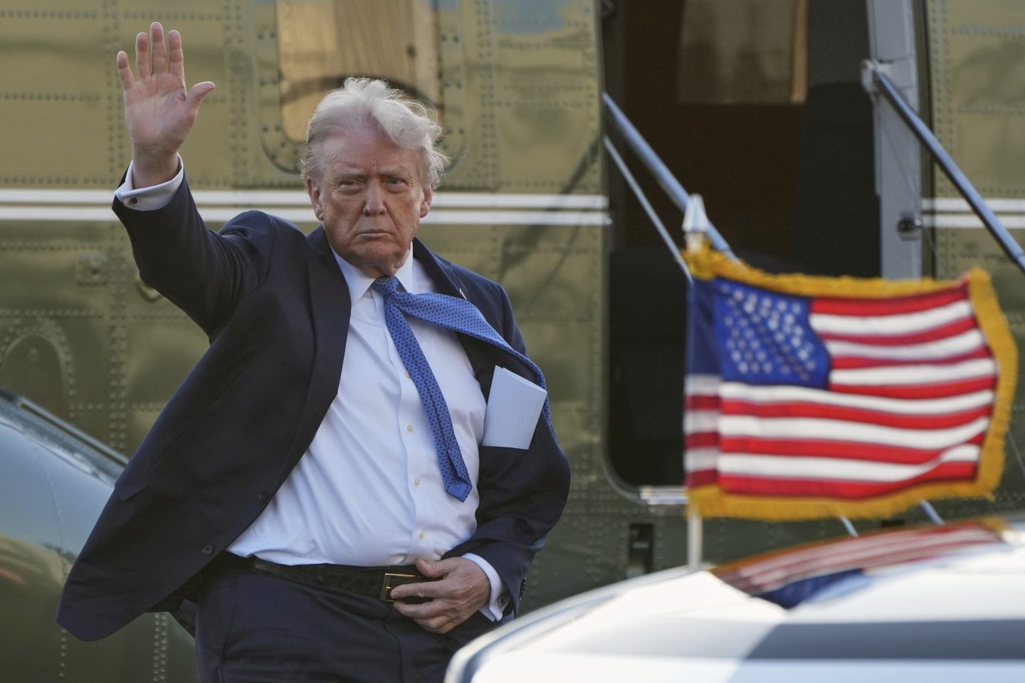 Trump waves to the press as a US flag waves on a car in front of him