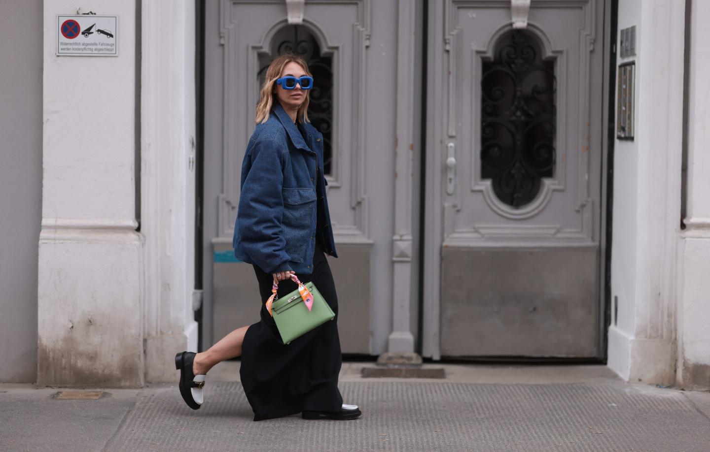 Karin Teigl seen wearing Loewe blue sunglasses, Louis Vuitton dark blue denim logo print pattern jacket, gold / silver jewelry, Saint Laurent black cotton shirt, Zara black fabric long skirt, Louis Vuitton black / white leather loafer, Hermès light green leather Kelly bag with a